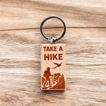 Laser engraved take a hike wooden keychain resting on wood surface, outdoor hiking themed wood keychain for camping gift, travel souvenir, or nature lover