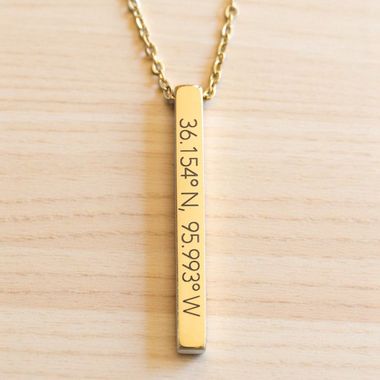 Vertical gold bar necklace engraved with Tulsa coordinates 36.154°N, 95.993°W, shown on a light wood background.
