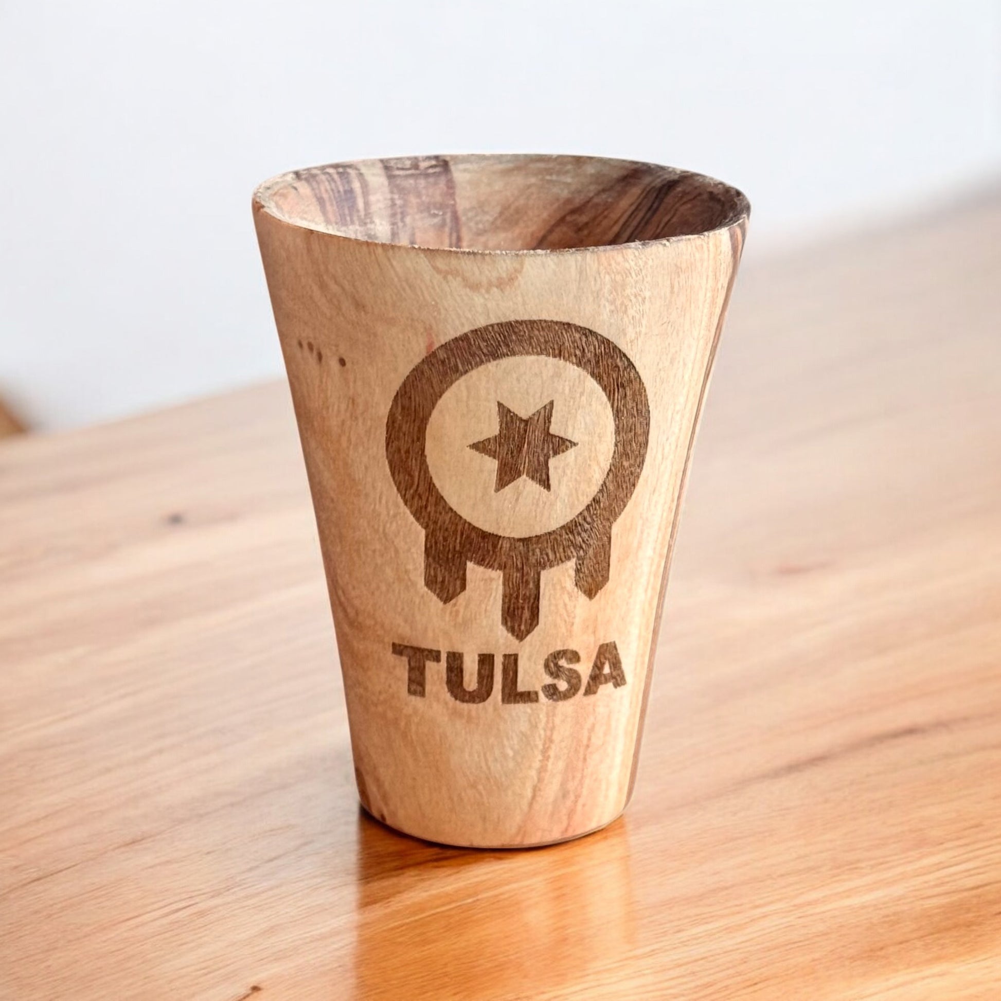 Olive wood shot glass engraved with Tulsa flag symbol shown on wood surface, Tulsa travel souvenir shot glass