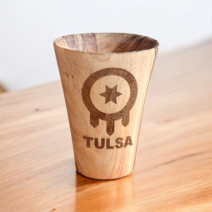 Olive wood shot glass engraved with Tulsa flag symbol shown on wood surface, Tulsa travel souvenir shot glass