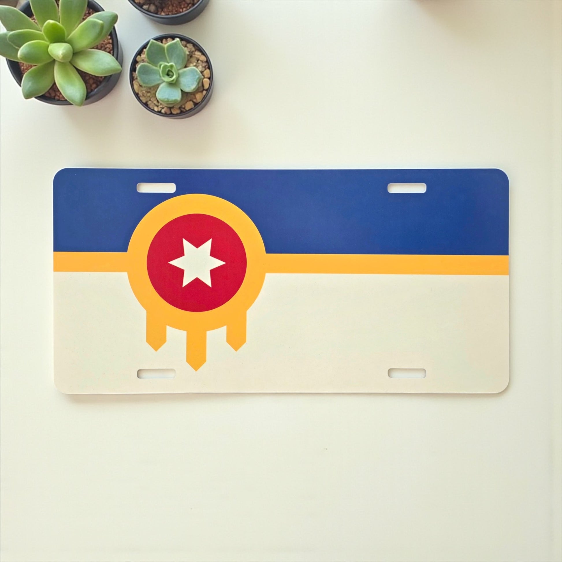 Tulsa Flag vanity license plate displayed on a white table with succulents, colorful Oklahoma souvenir by The Curious Bison.