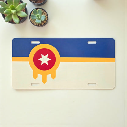 Tulsa Flag vanity license plate displayed on a white table with succulents, colorful Oklahoma souvenir by The Curious Bison.