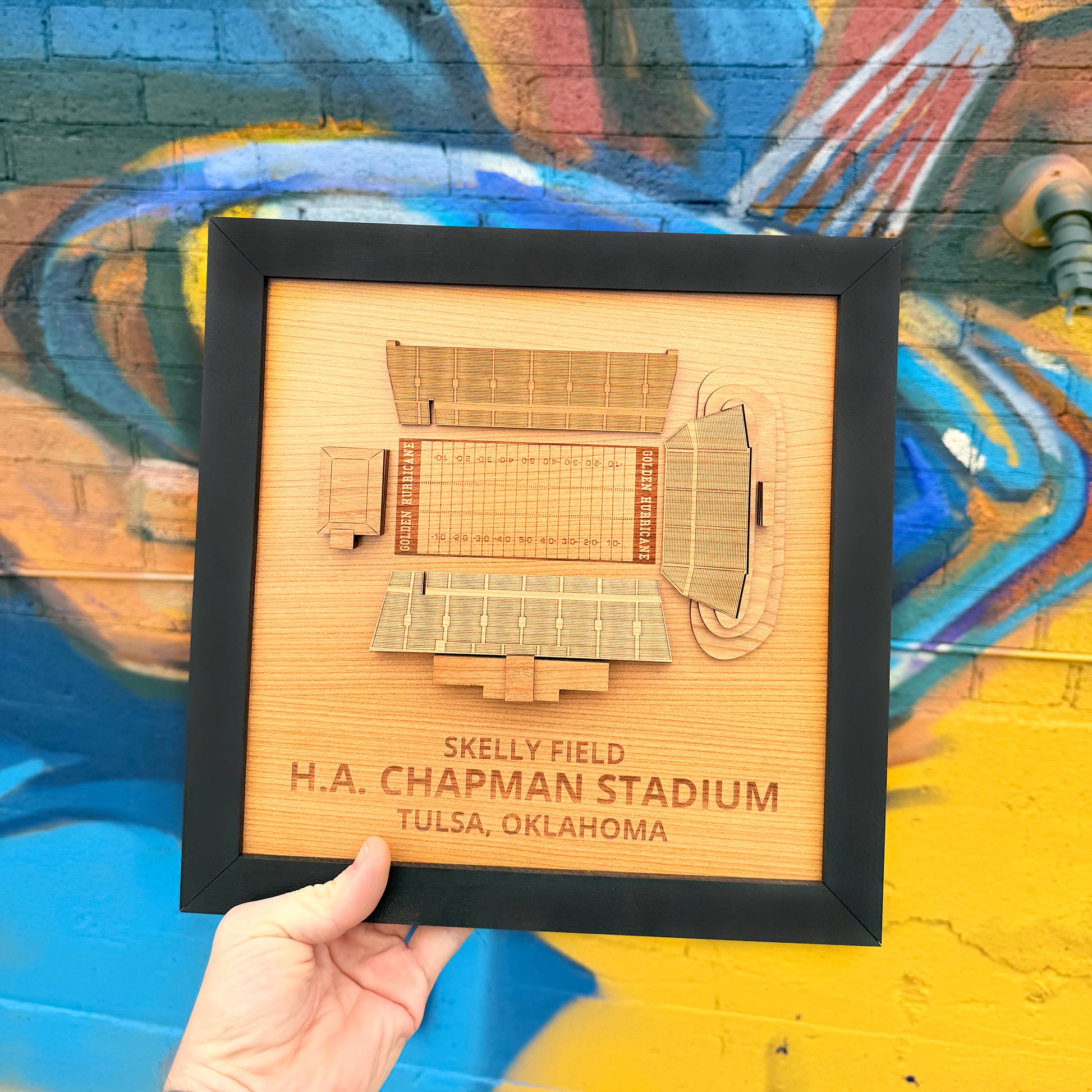 Hand holding framed Skelly Field H.A. Chapman Stadium layered wood wall art in front of colorful Tulsa mural creating Oklahoma souvenir and Tulsa football fan gift display