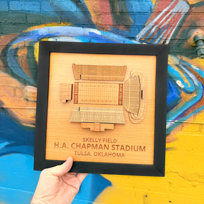 Hand holding framed Skelly Field H.A. Chapman Stadium layered wood wall art in front of colorful Tulsa mural creating Oklahoma souvenir and Tulsa football fan gift display