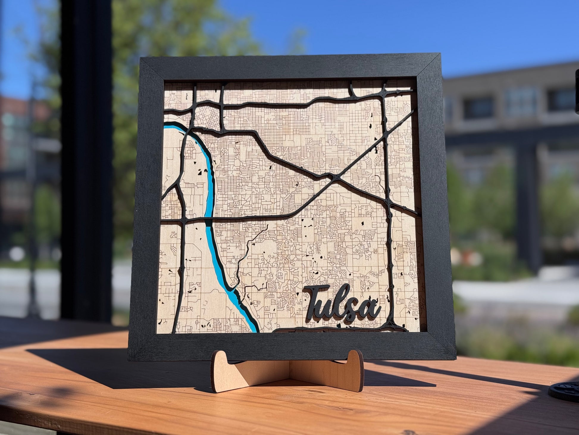 Framed Tulsa wooden city map displayed outdoors in natural light, layered laser cut wood map highlighting Tulsa streets and river as local wall art