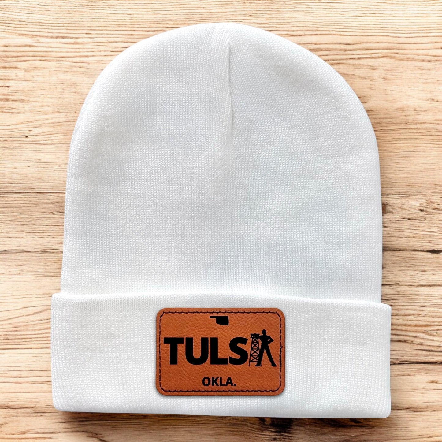 White Tulsa Oklahoma knit beanie laid flat on wood background with leatherette driller patch centered on cuff