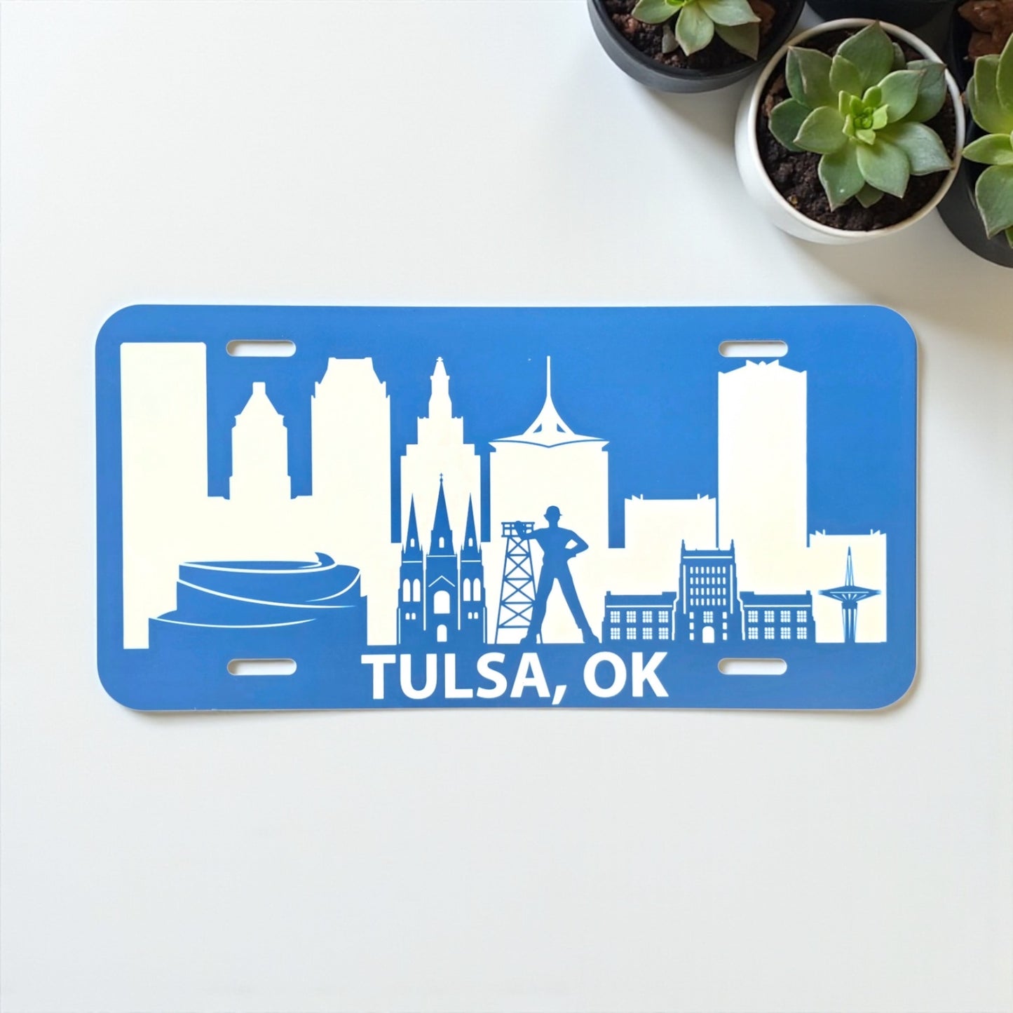 Blue Tulsa Skyline vanity license plate shown on a white background with potted plants, Route 66 souvenir by The Curious Bison.
