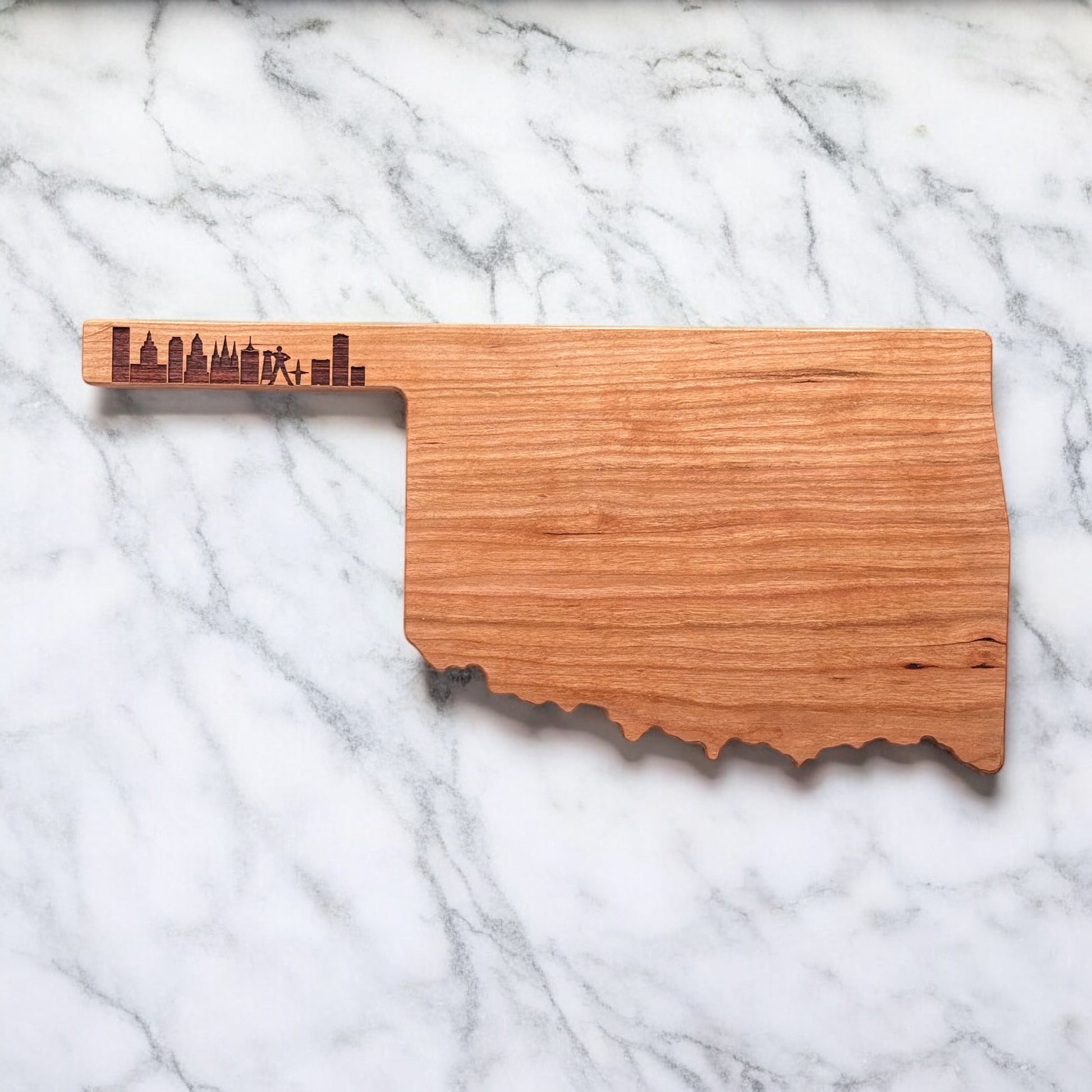 Flatlay of Oklahoma-shaped cherry charcuterie board with Tulsa skyline engraved handle on marble background.