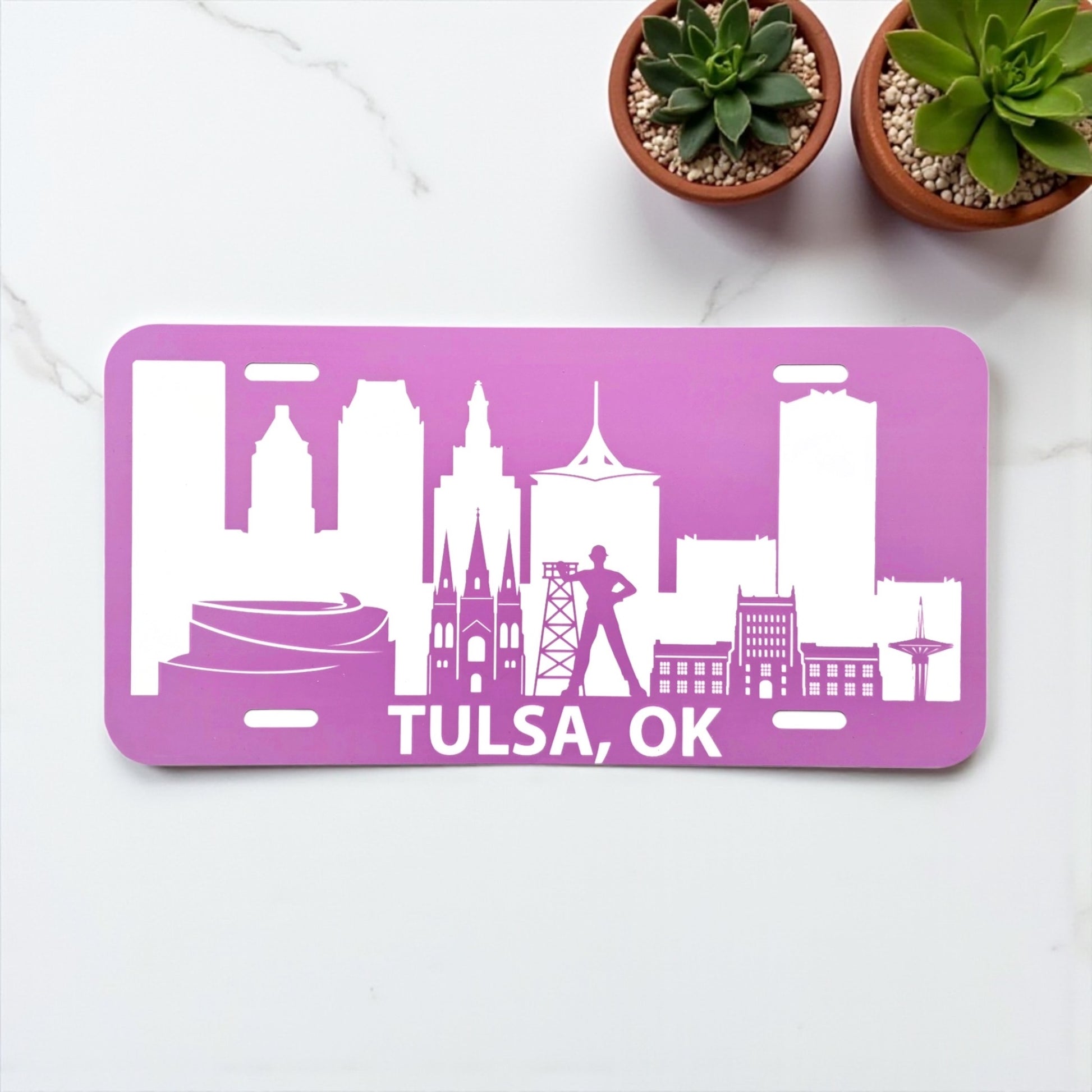 Pink Tulsa Skyline vanity license plate displayed on a white table with succulents, Oklahoma souvenir by The Curious Bison.