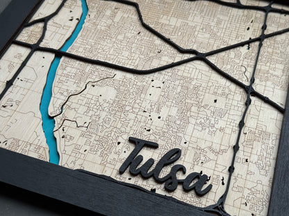 Close up of Tulsa name cut from black wood on layered wooden city map, engraved street detail visible in natural wood background