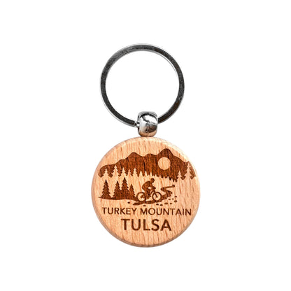 Round Turkey Mountain Tulsa wooden keychain with laser engraved cyclist on trail, trees, hills, and sun, lightweight wood keychain with metal ring for Tulsa souvenir or outdoor gift made in the USA
