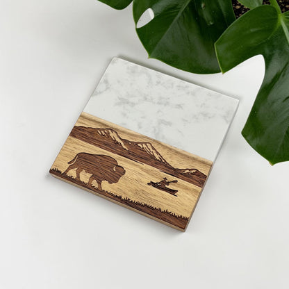 Marble and wood engraved coaster with mountain and bison artwork displayed beside green plant leaves, nature inspired coaster for rustic home decor and outdoor themed gifts