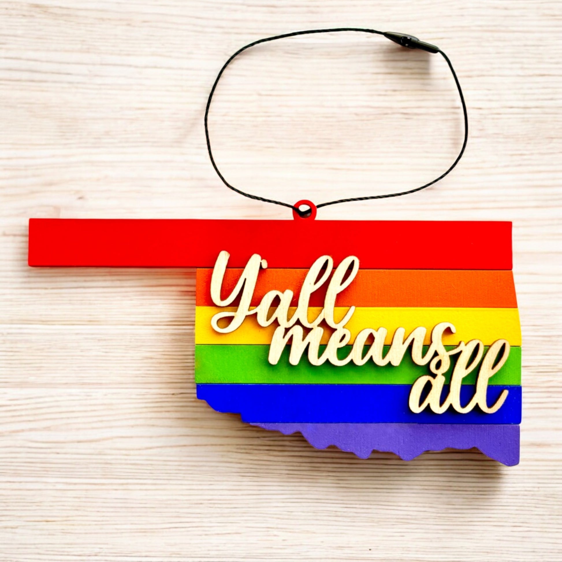 Flat lay of Y’all Means All Oklahoma rainbow wooden ornament showing layered pride colors, state shaped inclusive Oklahoma ornament