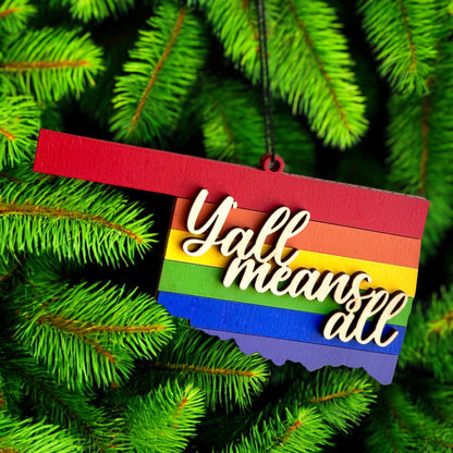 Y’all Means All Oklahoma rainbow ornament hanging on evergreen tree, layered wooden pride ornament used as holiday or year round decor