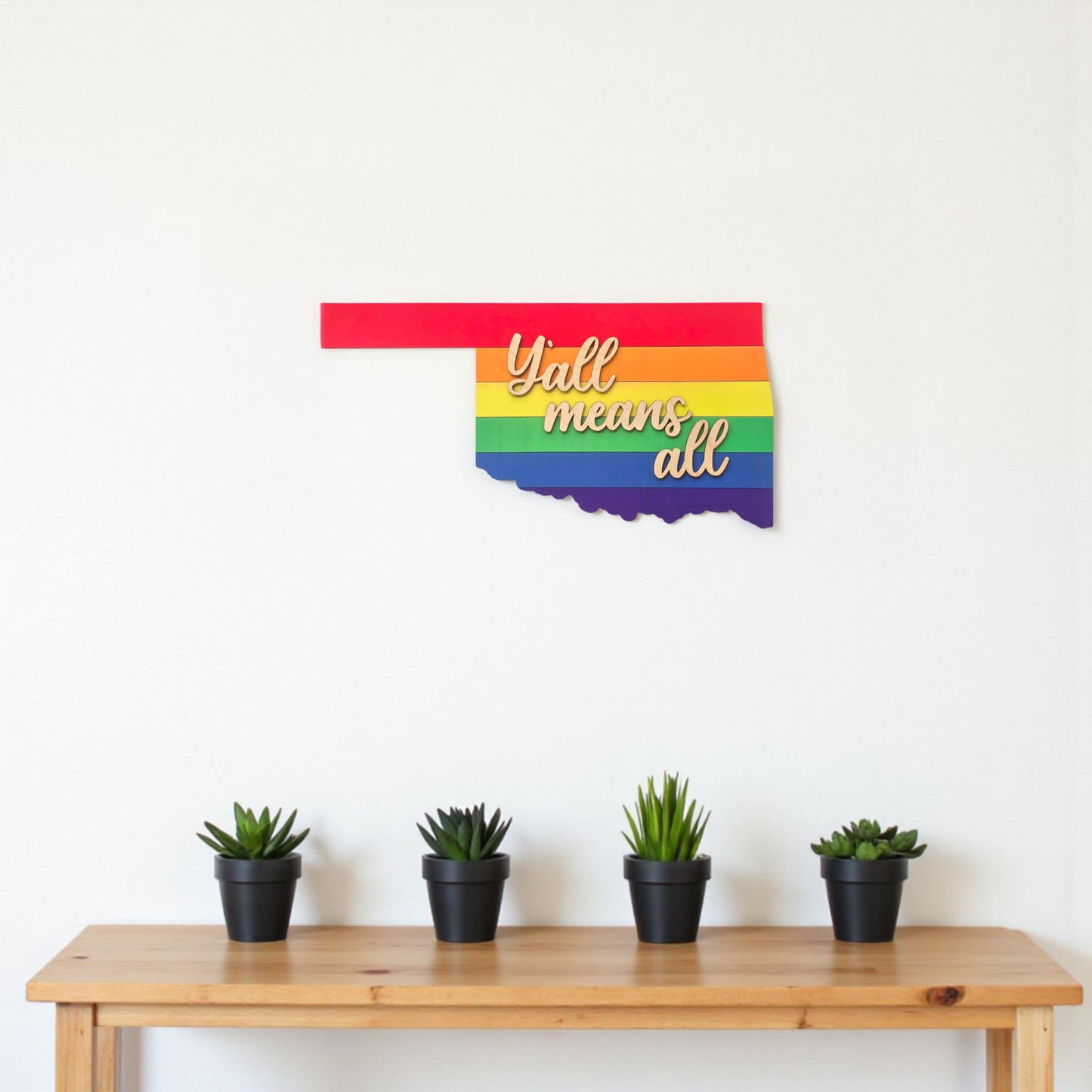 Oklahoma shaped wooden wall art with rainbow stripes and raised Y’all means all lettering displayed above a wooden shelf